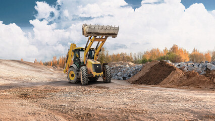 Fototapeta premium A loader lifts material at a construction site surrounded by piles of earth and rocks with trees in the background during daylight hours