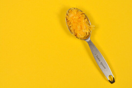 A top view of a stainless steel measuring tablespoon filled with fresh yellow lemon zest placed on a matching yellow surface with copy space