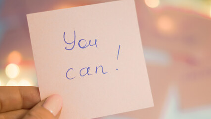 Hand holding pink sticky note with motivational message on blurred background