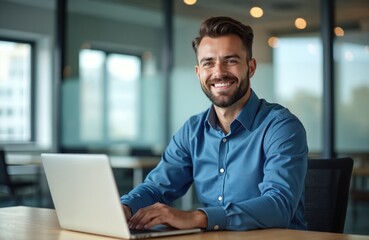 Fototapeta premium Young man in blue shirt sits at desk with laptop, smiles at camera. He works in modern office, looking happy and successful. Person uses computer, types on keyboard, focused. Business pro at work.