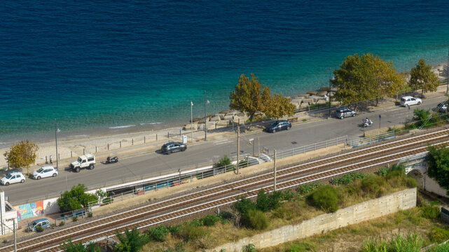 High-angle aerial view of a coastal road with parked cars, a double-track railway, and a beach with turquoise water. It is an italian coastline road on a summer sunny morning.