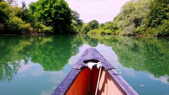 First-person view shows the tip of a canoe gliding along a calm river, with green vegetation and trees reflected on the water.