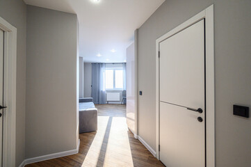 Fototapeta premium minimalist hallway with light grey walls, white doors, wooden flooring, recessed ceiling lights; opens to a bright living area with large window and sheer curtains.