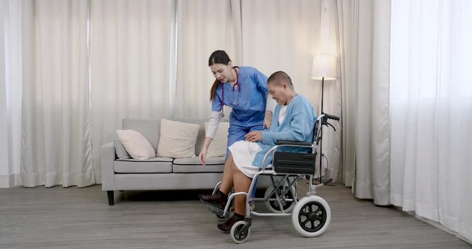 Asian female nurse pushing senior female cancer survivor in wheelchair across living room during recovery phase highlighting homecare assistance and mobility support in healing journey