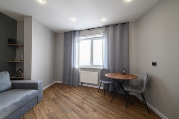 Fototapeta premium cozy modern room with a grey sofa, wooden flooring, a round dining table with two chairs, grey curtains, built-in shelves, recessed ceiling lights, and a large window with natural light.