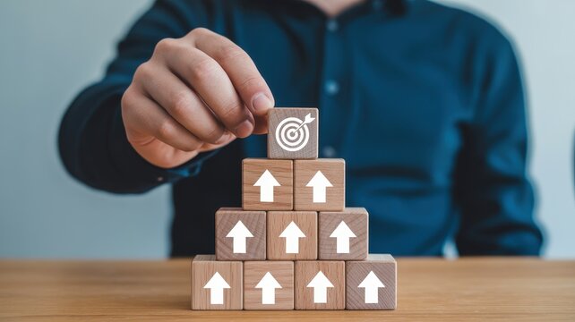 Hand placing a wooden block with a target icon on a pyramid of blocks with upward arrows, representing business goals and growth.