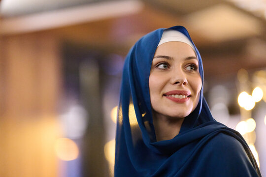 Smiling Woman in Blue Hijab Outdoors at Night With Warm City Lights Behind