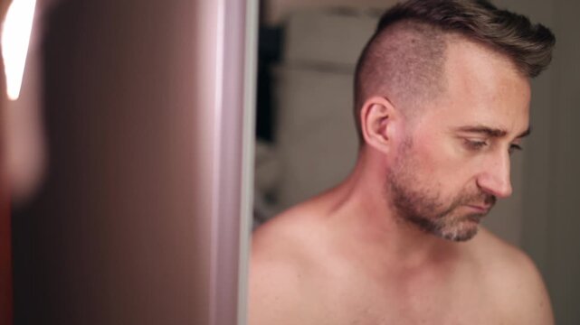 Man grooms himself, Man reflects on grooming in mirror, Gentle man scrutinizes stubble in reflective bathroom setting, Caucasian man thoughtfully examines facial stubble in mirror with soft lighting
