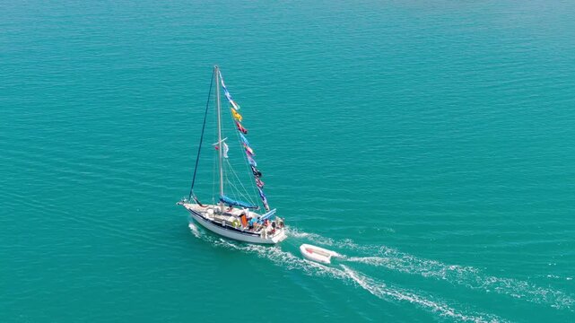 High-angle aerial drone video following a sailing yacht with flags, towing a dinghy across a vibrant turquoise ocean