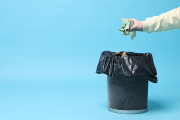 Woman throwing napkin into trash bin on light blue background, closeup. Space for text