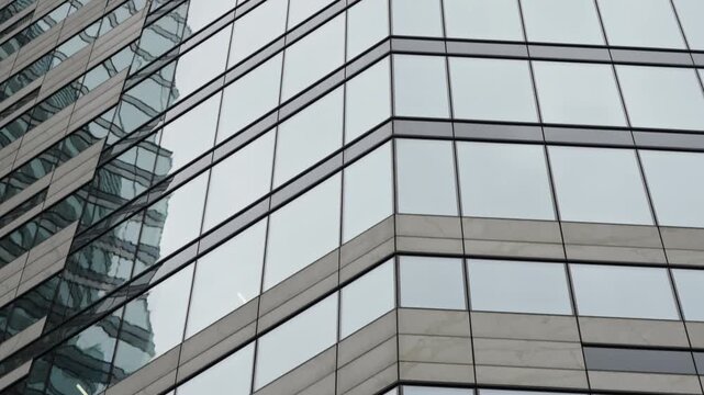 Modern glass-clad office tower facade with repeating window grid and sharp corner lines, reflecting neighboring high-rise forms across mirrored panels in an overcast urban skyline
