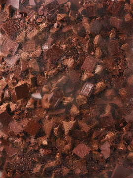 Assorted chocolate pieces are seen through wet glass with fine water droplets. Broken bars and chocolate pearls form a layered texture that enhances the rich cocoa colors