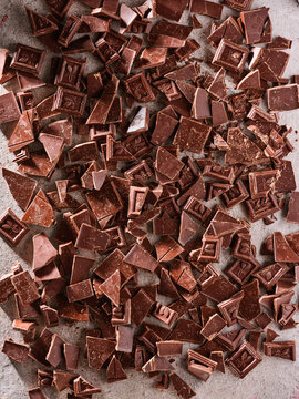 Small pieces of chocolate are scattered on a gray surface. These pieces are ready for use in baking and cooking. They vary in shape and size, showing some detail of the chocolate