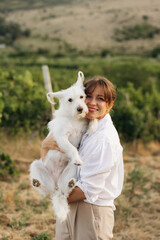 Naklejka premium Portrait of a smiling young woman with a white fluffy dog against a background of vineyards at sunset. Concept of nature, family, friendship, and harmony