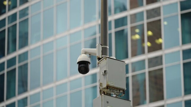 Pole-mounted dome surveillance camera with control box faces a reflective glass office facade, illustrating urban security infrastructure, safety, and modern monitoring systems.