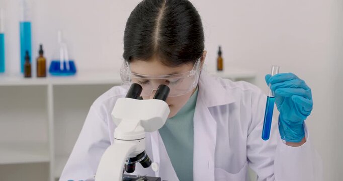 Closeup of asian schoolgirl focusing on microscope while wearing safety goggles and gloves during classroom science experiment with blue liquid in test tube in hand, learning scientific method