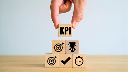 Hand placing wooden block with KPI text on top of stacked cubes with business icons, representing performance measurement and business success