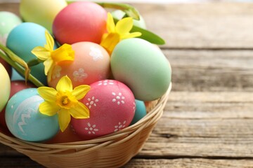 Fototapeta premium Painted Easter eggs and beautiful daffodils in wicker basket on wooden table, closeup. Space for text