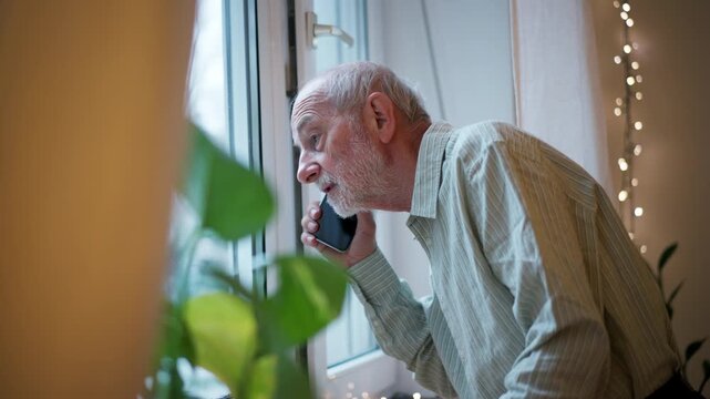 Old man standing by window, making phone call.