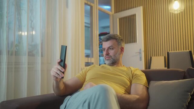 Handsome man using remote controller while watching tv on couch.