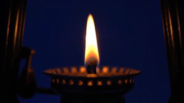 A sequence of multiple small oil lamps burning with steady, bright flames against a deep navy background. The atmospheric light flickers gently across the metallic holders, emphasizing warmth and dar