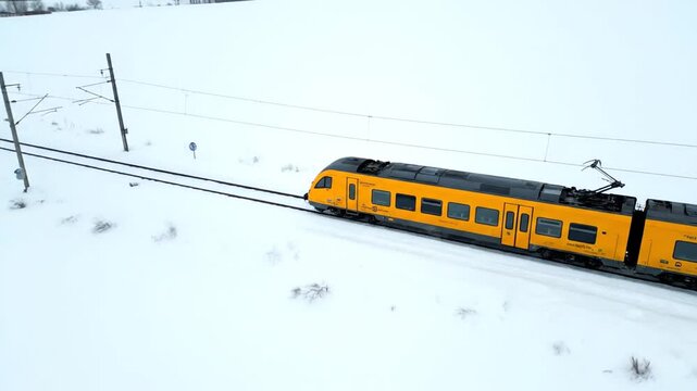 A bright yellow electric train speeds across snow-covered fields on parallel tracks. The high-angle view captures the train traveling through the stark winter landscape under overhead power lines.