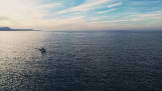 Drone aerial 4k resolution footage of fishing boat sailing away on calm ocean water with a mountain range on the horizon under a soft sunset or sunrise sky. Akamas Paphos Cyprus