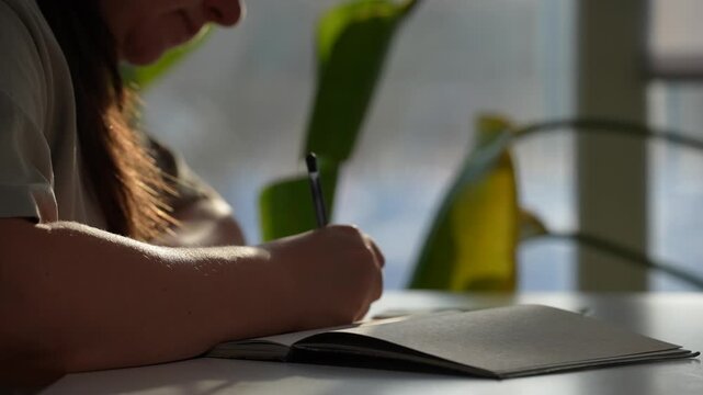 Woman writing with pen in notebook on white table with window background