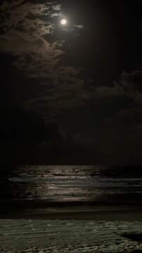 The moon is shining brightly in the dark sky over the ocean. The water is calm and the sky is filled with clouds. The scene is peaceful and serene, with the moonlight reflecting off the water