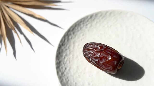 A single brown date slowly rotates on a textured white plate. The minimalist composition features harsh light and dramatic shadows cast by a dried palm frond, highlighting the fruit's texture and ric