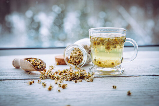 Matricaria chamomilla, commonly known as chamomile, wild chamomile, or scented mayweed. Organic dried herbal tea powder made from flower blossoms. Tea in a glass mug on a wooden windowsill, with snowy
