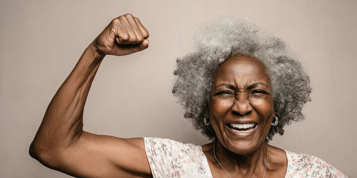 Empowered senior Black woman with grey hair flexing her bicep and laughing, representing health, strength, and vitality in aging.