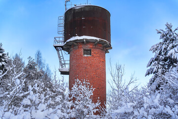 Old red brick water tower in snowy winter forest.Vintage red brick water tower surrounded by snow-covered trees under a bright blue winter sky
