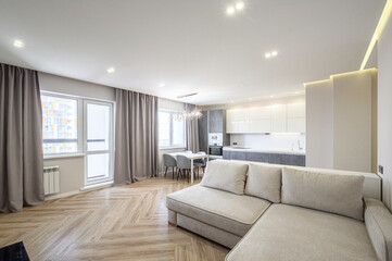 Spacious modern living room with beige sofa, herringbone wood flooring, dining area, kitchenette, large windows with grey curtains, recessed ceiling lights, chandelier.