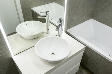 Fototapeta premium Modern bathroom with two round vessel sinks, chrome faucets, white countertop, LED-lit mirror, grey marble tiles, sleek bathtub, minimalist design.