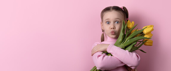 Cheerful young girl with pigtails hugs a bouquet of yellow tulips against a pastel pink background. Copy space for spring or celebration themes.
