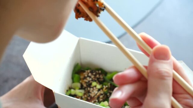 Person hands holding paper box takeout food, skillfully using wooden chopsticks pick up piece of stir fried shrimp, with sesame seeds, green onions. Woman eating asian shrimp food with chopsticks