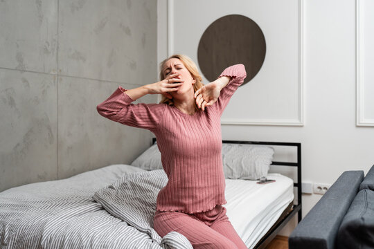 A young woman in pajamas yawns and stretches while sitting on a bed in a modern bedroom at home.