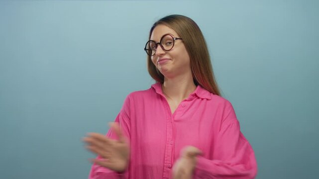 Young woman with glasses in pink shirt stands over isolated blue background, expressing boredom and indifference with subtle gestures and facial expressions.