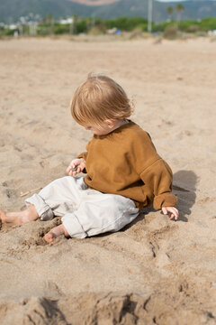 Toddler sitting on a sandy beach and exploring sand with hands. Natural daylight, soft focus background, and warm neutral colors create a calm lifestyle scene. Concept of childhood, curiosity, sensory
