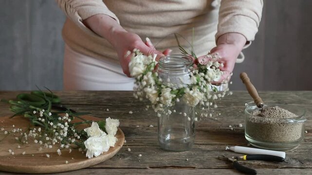 Florist at work: woman shows how to make romantic candle holder using a glass jar, carnations flowers, gypsophila paniculata twigs and sand. Step by step, tutorial. Images available.
