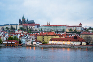 Obraz premium A scenic Prague riverside view showing red-roofed buildings, a grand historic castle, and bustling waterfront venues along the Vltava. Ideal for travel, culture, and European city life themes.
