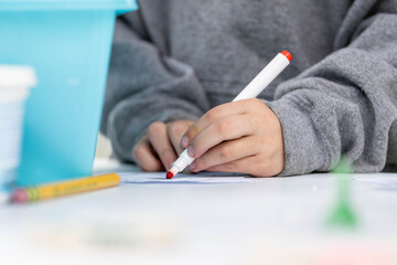 A student's hand grips a red marker over white paper at a desk, blurred classroom behind