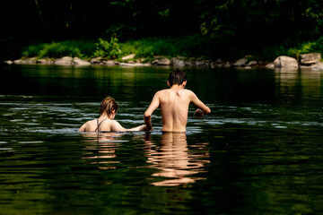 Fototapeta premium Back view of two people wading and swimming in deep natural river or lake with dark green water and rocky forested shoreline. 