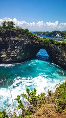 Arch of rock over vibrant, churning turquoise sea with jagged cliffs on the horizon under a bright blue, cloud-dappled sky
