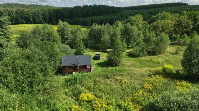 Aerial sequence descends toward a small wooden cabin, stainless chimney, dark car, and yellow wildflowers in a birch and mixed forest under midday summer light.