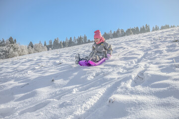 A joyful child sledging on a pink sledge, surrounded by a picturesque winter landscape, showing pure delight while enjoying the snow on a bright sunny day