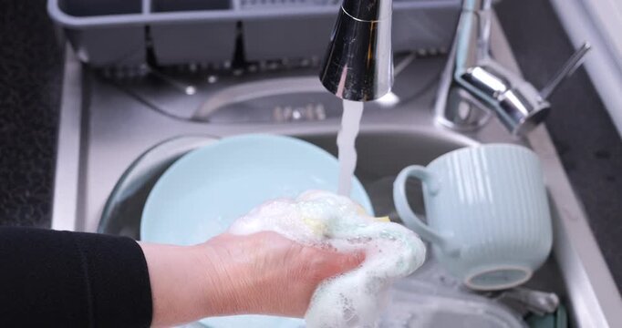 Close-up of a hand squeezing a sponge full of foam under running water in a sink full of dishes. Dish washing detergent, cleaning power and household hygiene concept.