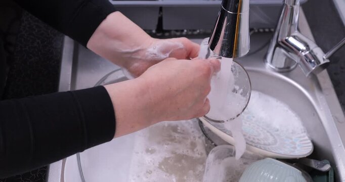 Close-up of rinsing a mug covered in foam under running water in a sink full of dishes. Dish washing, cleanliness and detergent effectiveness concept. Slow motion