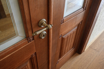 Obraz premium A pair of wooden interior doors with glass panels, ornate brass handle and keyhole, rich brown finish, decorative corner carvings, set against light wooden flooring.